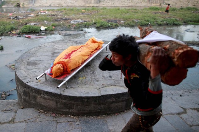Labourer carrying wood walks past the wrapped body of a victim of Saturday's earthquake before its cremation along a river in Kathmandu, Nepa