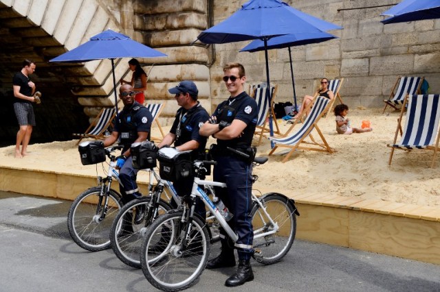 Policemen stand on their bikes as people relax under parasols in "Paris Plage" (Paris Beach) during the opening day of the event on the bank of the Seine river, in central Paris, on July 20, 2016. The 15th edition of Paris Plage will run until September 4, 2016. / AFP PHOTO / BERTRAND GUAY