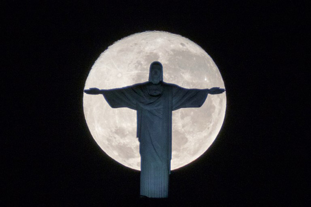 The silhouette of the statue of Christ the Redeemer atop Corcovado hill stands out against the full moon in Rio de Janeiro, Brazil, on July 19, 2016. / AFP PHOTO / YASUYOSHI CHIBA