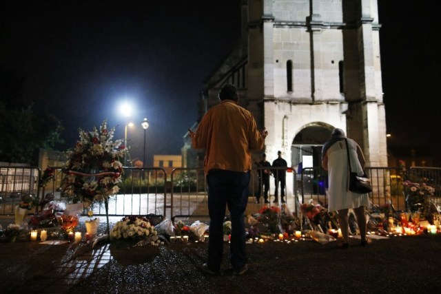 People stand in front of a makeshif memorial near two gendarmes guarding the Saint-Etienne du Rouvray church on July 27, 2016, after the priest Jacques Hamel was killed on July 26 in the church during a hostage-taking claimed by Islamic State group. France probes an attack on a church in which two men described by the Islamic State group as its "soldiers" slit the throat of a priest. An elderly priest had his throat slit in a church in northern France on July 26 after two men stormed the building and took hostages. The attack in the Normandy town of Saint-Etienne-du-Rouvray came as France was still coming to terms with the Bastille Day killings in Nice claimed by the Islamic State group. / AFP PHOTO / CHARLY TRIBALLEAU