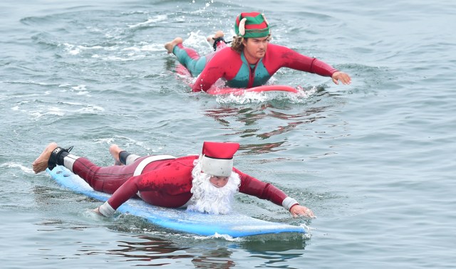 Surfing Santa, Michael Pless, and Jonathan Livingston, dressed as an Elf, make their move to catch a wave at Seal Beach, California on December 10, 2016, where he runs a surfing school and has every December since in 1990's gone out to surf in his Santa Claus outfit. / AFP PHOTO / Frederic J. BROWN