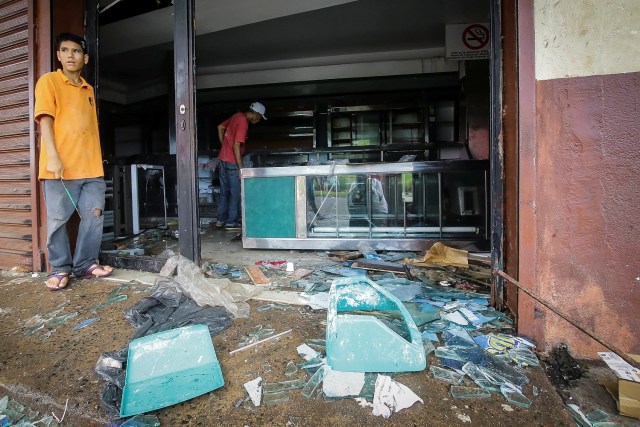 People stand next to broken glass as they look at the damage after a bakery was looted in Ciudad Bolivar