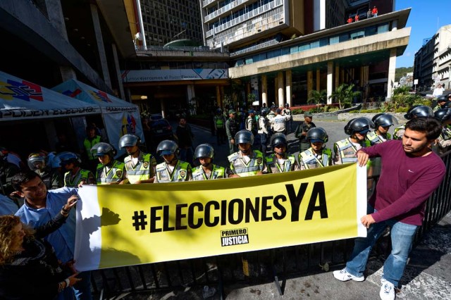 Riot police stand behind a banner that reads "Elections now" in fornt the National Electoral Council (CNE) in Caracas on January 2, 2017. A group of opposition lawmakers had their way blocked by police when they tried to get into the CNE to demand the organism to call for regional elections that were due to happen last year. / AFP PHOTO / FEDERICO PARRA