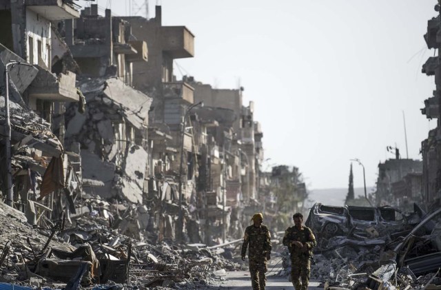 Fighters of the Syrian Democratic Forces (SDF) walk down a street in Raqa past destroyed vehicles and heavily damaged buildings on October 20, 2017, after a Kurdish-led force expelled Islamic State (IS) group fighters from the northern Syrian city, formerly their "capital". SDF fighters flushed jihadist holdouts from Raqa's main hospital and municipal stadium, wrapping up a more than four-month offensive against what used to be the inner sanctum of IS's self-proclaimed "caliphate", which for three years saw some of the group's worst abuses and grew into a centre for both its potent propaganda machine and its unprecedented experiment in jihadist statehood. / AFP PHOTO / BULENT KILIC