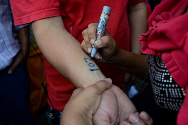A woman writes numbers on people's arms so they know what order they came in, as they queue outside a supermarket in Caracas to buy basic foodstuffs and household products on November 10, 2017. In crisis-stricken Venezuela, the cost of the basic basket of goods soared to nearly 2.7 million bolivars in September, the equivalent of six minimum monthly wages. / AFP PHOTO / FEDERICO PARRA