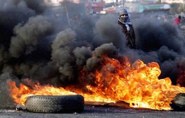 A Palestinian protester walks with slingshot through the smoke of burning tires during clashes with Israeli forces in the West Bank city of Ramallah on December 11, 2017 as demonstrations continue to flare in the Middle East and elsewhere over US President Donald Trump's declaration of Jerusalem as Israel's capital. / AFP PHOTO / THOMAS COEX