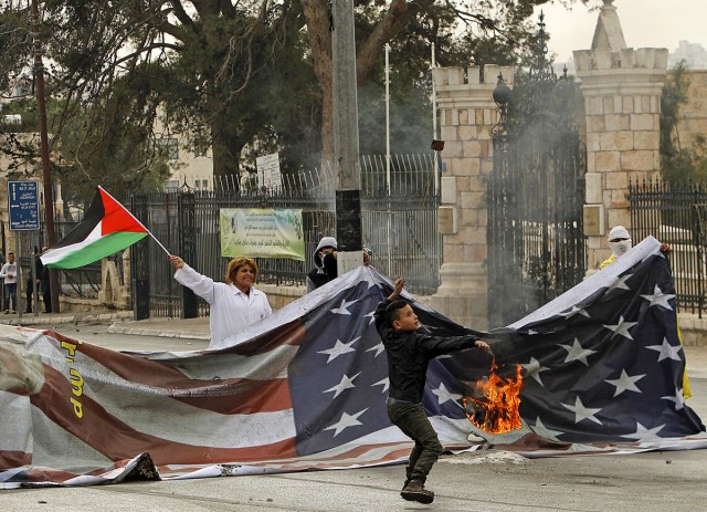 A Palestinian youth uses a slingshot to hurl stones in front of a burning banner bearing the US flag, while a woman waves a Palestinian flag behind, during clashes with Israeli security forces at the main entrance of the occupied West Bank city of Bethlehem on December 20, 2017 as protests continue following the US president's controversial recognition of Jerusalem as Israel's capital. / AFP PHOTO / Musa AL SHAER
