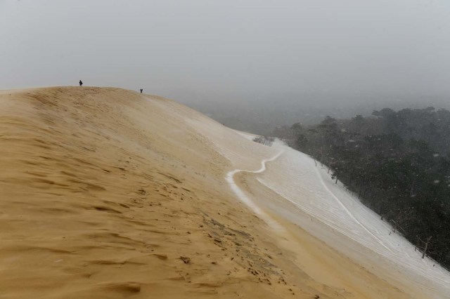 People walk on the partially snow covered Pilat (or Pyla) sand dune during snow fall on February 28, 2018 in La Teste-de-Buch, southwestern France. Europe remained on February 28 gripped by a blast of Siberian weather, accounting for at least 24 deaths and carpeting palm-lined Mediterranean beaches in snow. / AFP PHOTO / NICOLAS TUCAT