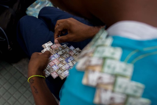 Wilmer Rojas, 25, sists at a bus stop in Caracas as he sews Bolivar bills, to make a paper crown on January 30, 2018. A young Venezuelan tries to make a living out of devalued Bolivar banknotes by making crafts with them. / AFP PHOTO / FEDERICO PARRA