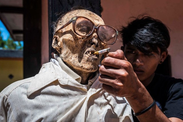 PIC BY Claudio Sieber / CATERS NEWS - (PICTURED A relative lights a cigarette) THESE images show a unique insight into the village where the living dig up their dead relatives to clean their bones and to dress them up. The dead can even remain in homes for decades until the family throw a huge party in their honour. The corpses are treated as sick until they finally find peace after funeral rites known as Rambu Solo The bizarre ritual was pictured in South Sulawesi, Indonesia, where even buried relatives are taken out of their coffins for new outfits and a bone polish. SEE CATERS COPY