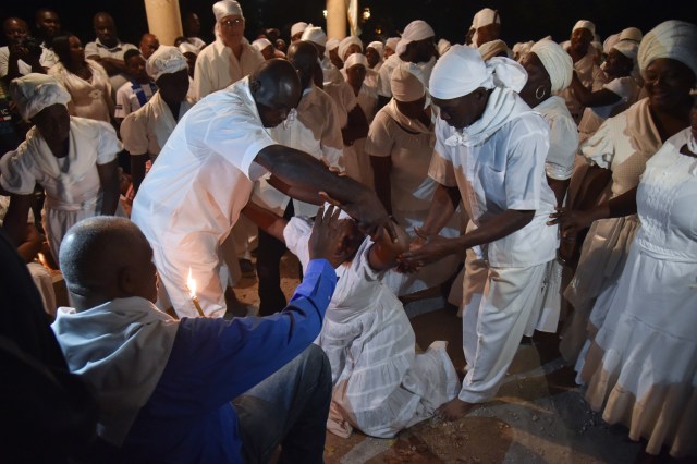 A Haitian woman kneels on the ground supported by fellow voodoo followers wearing white clothes, as they dance while participating in a voodoo ceremony in Souvenance, a suburb of Gonaives, 171km north of Port-au-Prince, on March 31, 2018. Haitian voodoo followers arrived in Souvenance to take part in the voodoo ceremonies held during Easter weekend. / AFP PHOTO / Hector RETAMAL