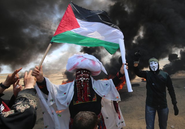Un palestino sosteniendo su bandera nacional camina en el humo de neumáticos quemados junto a un manifestante con una máscara anónima durante enfrentamientos con las fuerzas israelíes en la frontera con la franja de Gaza al este de Khan Yunis el 14 de mayo de 2018, mientras los palestinos protestan por la inauguración de la embajada de los Estados Unidos después de su controvertido traslado a Jerusalén. / AFP PHOTO / SAID KHATIB