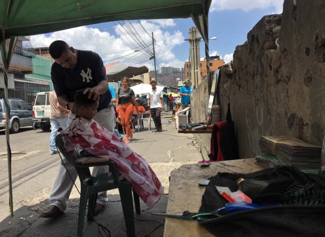 Un hombre corta el cabello de un niño en una calle de Caracas el 4 de junio de 2018. Bajo un puente o en un mercado al aire libre en Caracas, los peluqueros se ganan la vida con herramientas simples y ofrecen cortes de pelo para aquellos que no pueden pagar un salón de belleza debido a la crisis económica. / AFP PHOTO / Juan BARRETO